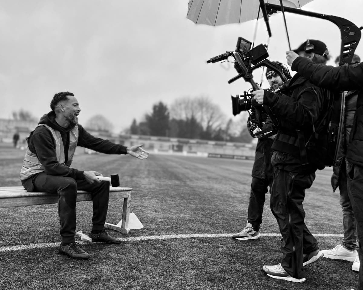 A man in a hi-vis vest sits on a bench on a football pitch, gesturing to a film crew with a camera rig and an umbrella