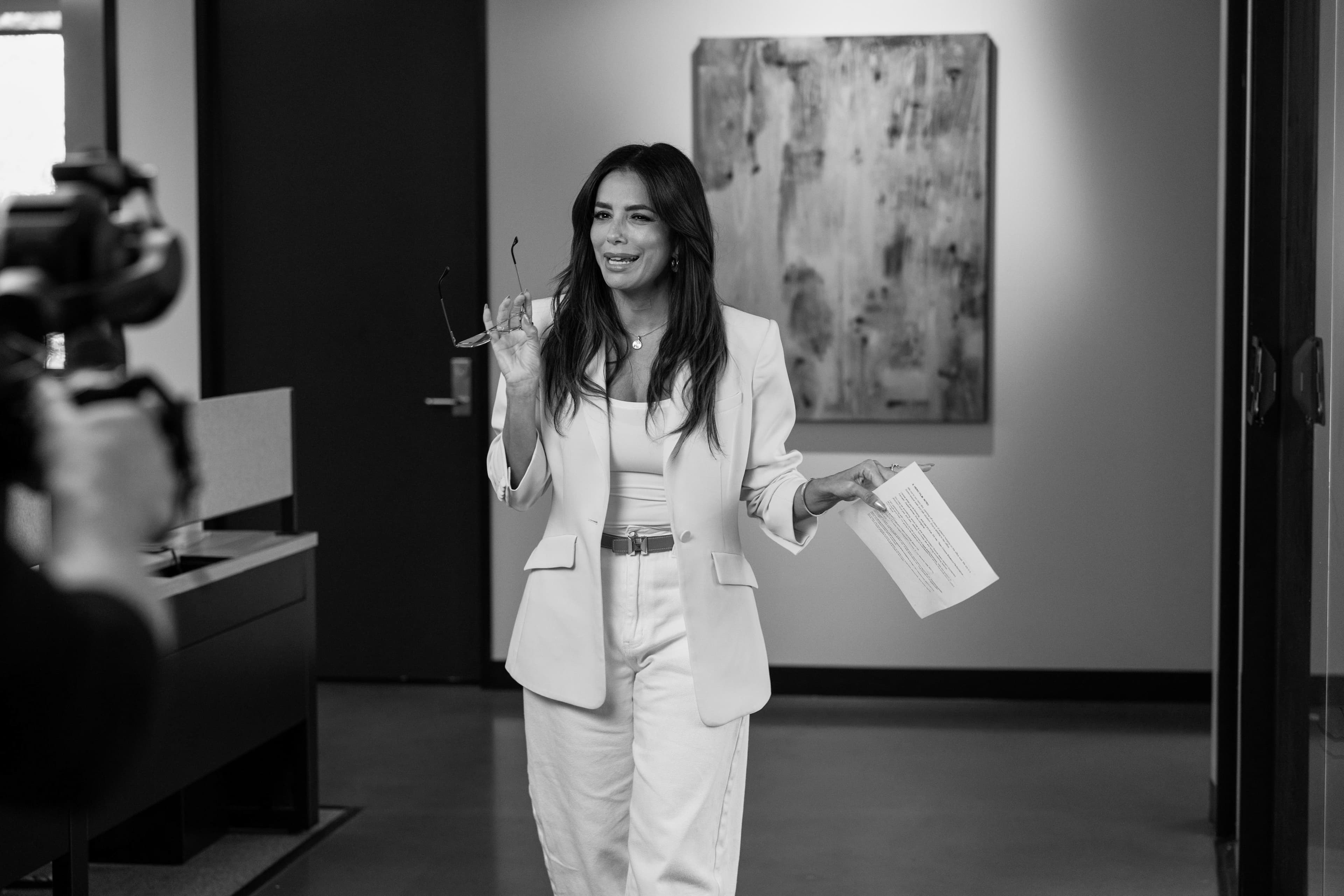 A woman in a white suit walks towards a film camera, glasses in one hand and a script in the other